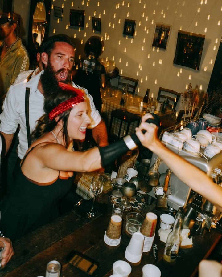 A guest in a red headband reaches across a bar toward another raised hand, with cups and warm reflections around them.