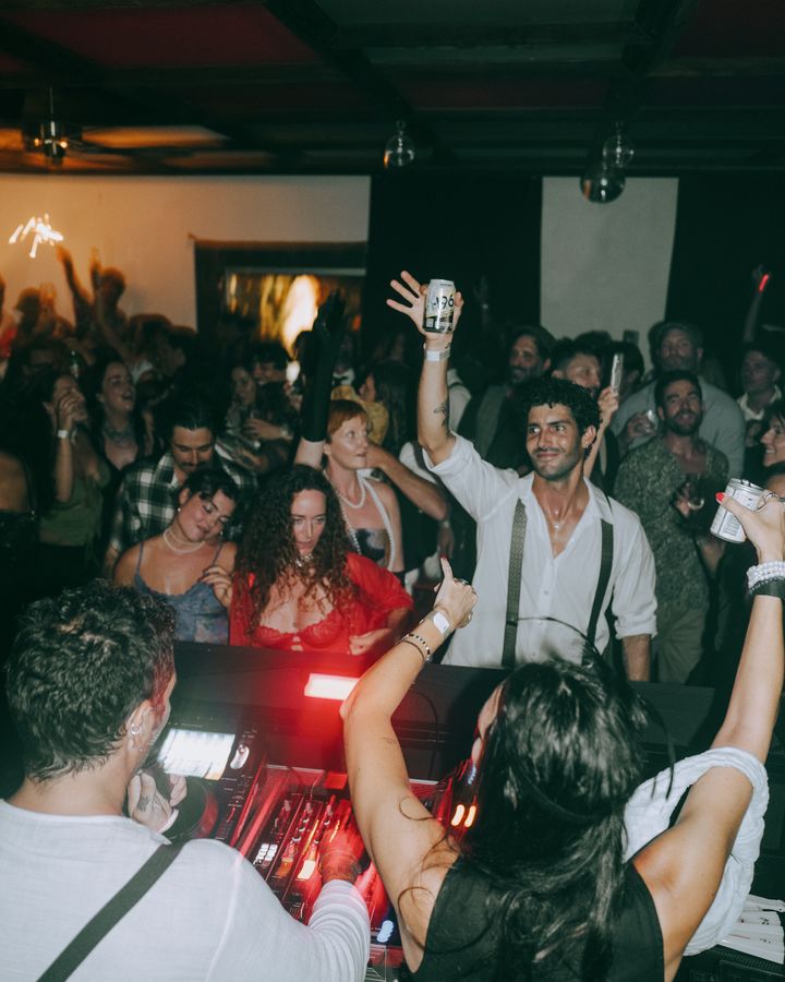 A packed dance floor seen from behind a mixing desk, with guests raising drinks and hands under red ceiling lights.
