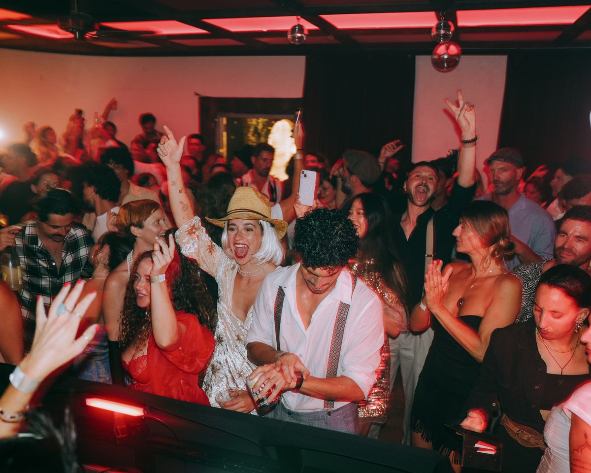 A wide crowd of guests dances with raised hands beneath red ceiling panels and mirrored balls, seen across the front edge of a DJ booth.