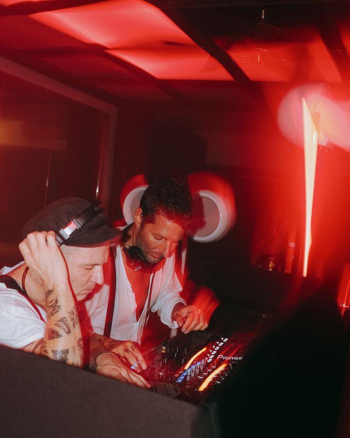 Two DJs in white shirts work at a mixing desk under red lights, one holding headphones to an ear.