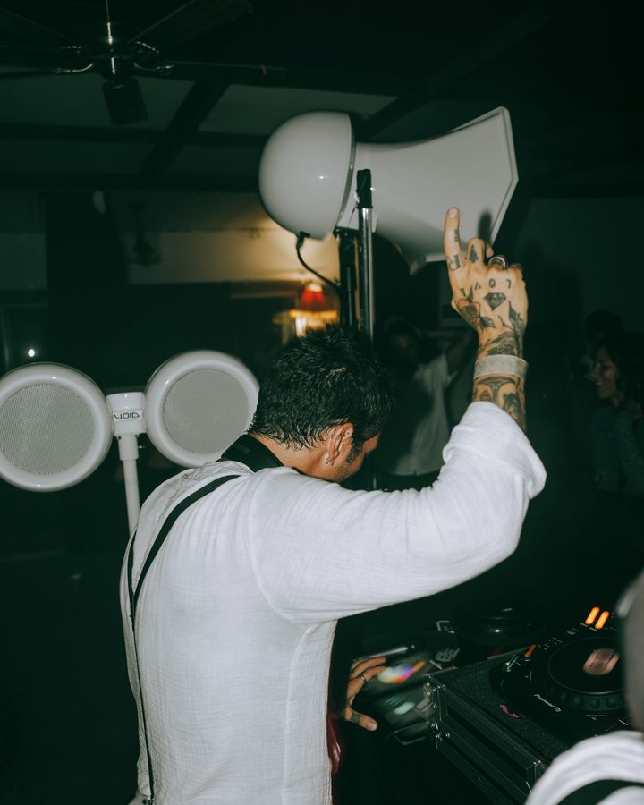 A DJ seen from behind raises one hand above the decks, beside large white speakers and a dimly lit crowd.