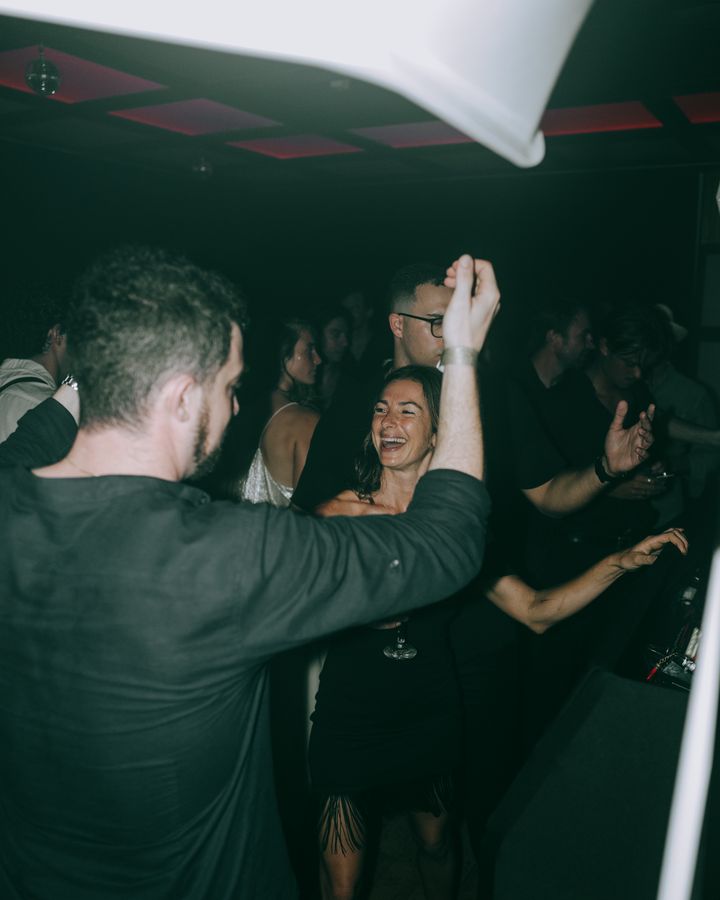 A smiling guest in black dances facing another guest with a raised arm, surrounded by people on the dance floor.