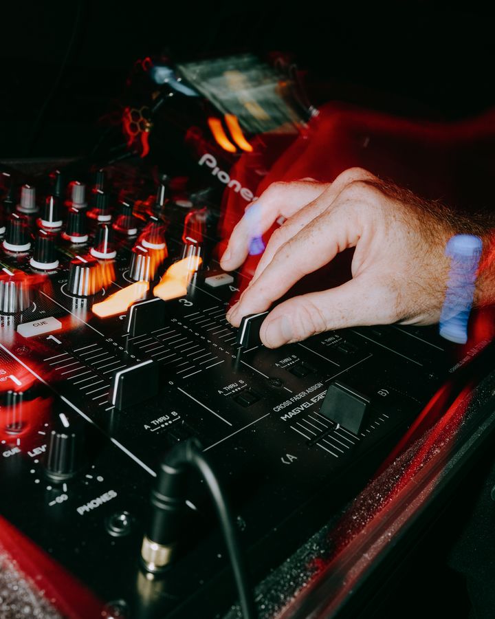 A close-up of a hand adjusting a fader on a DJ mixer, surrounded by controls and red and orange light trails.