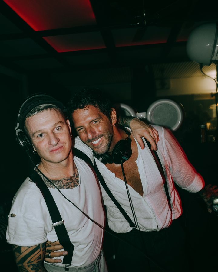 Two people in white shirts and braces pose with an arm around one another, wearing headphones beside white speakers.