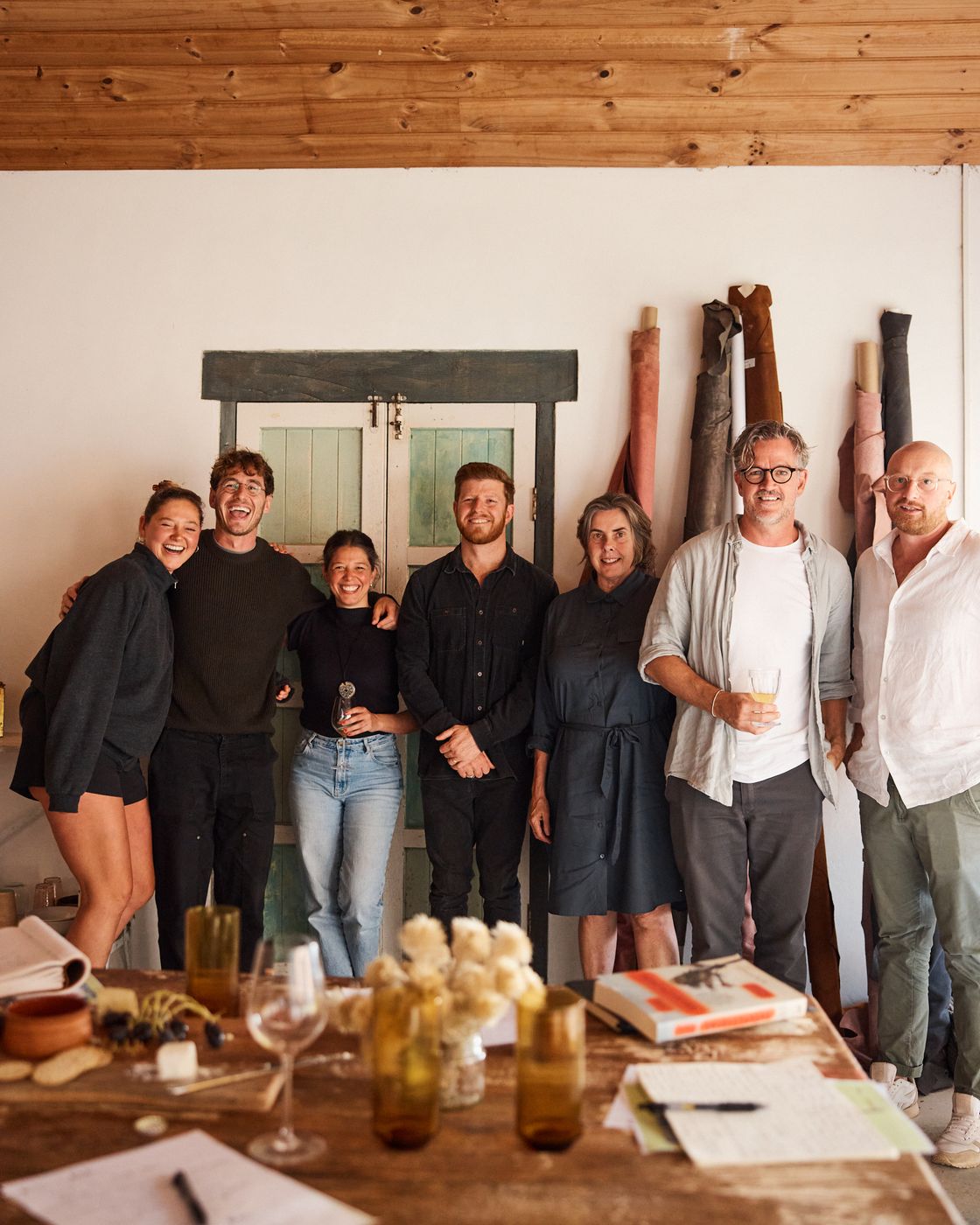 Seven people stand smiling together in front of an old green double door and rolls of fabric, beneath a timber ceiling, with the Bali table in the foreground.