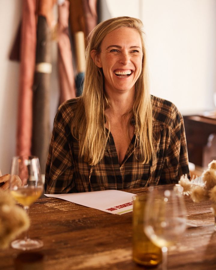 A woman in a checked shirt laughs at the Bali table, with wine glasses, dried flowers and a writing sheet in front of her.