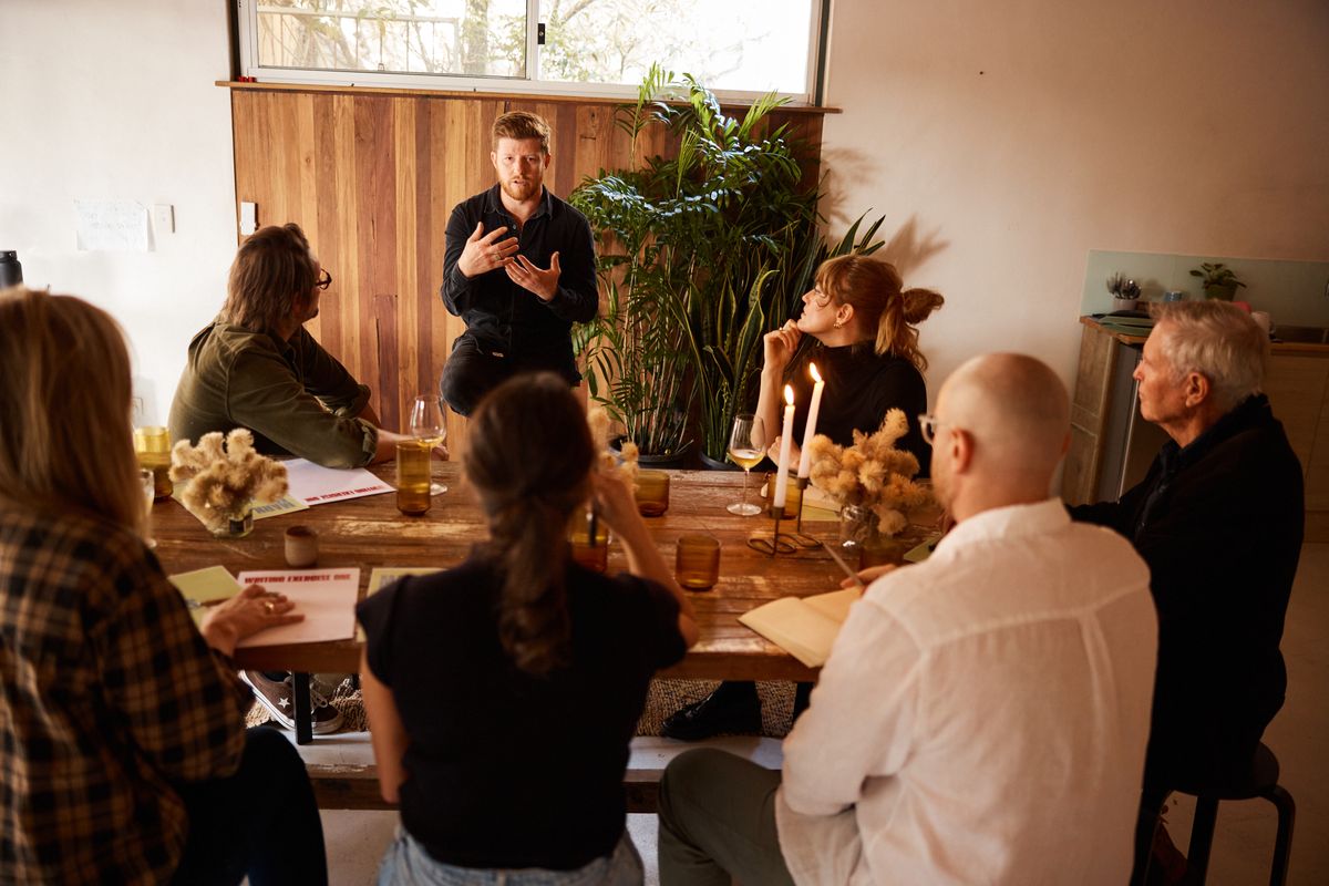 A man in a dark shirt talks with his hands to a group of writers around the Bali table, with candles, dried flowers and palms in the room.