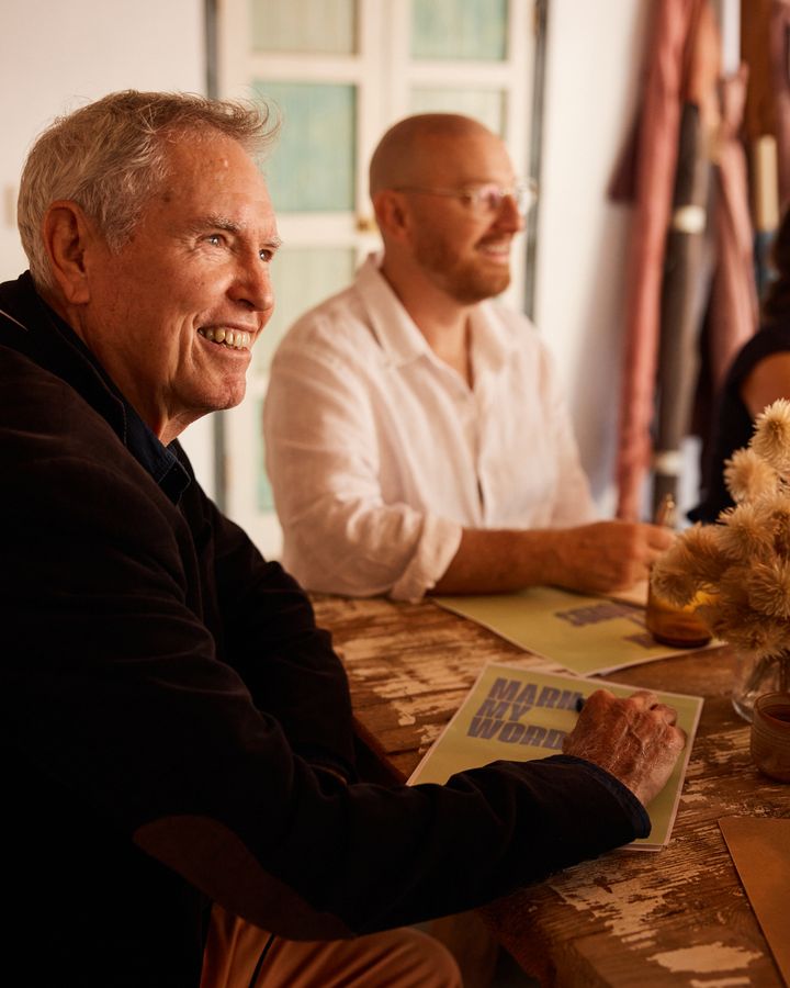 An older man smiles as he writes on a green Mark My Words booklet at the Bali table, with another writer in a white shirt beside him.
