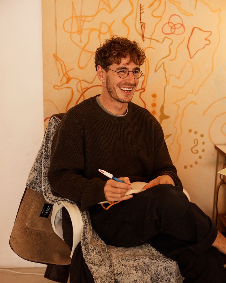 A man with curly hair and round glasses laughs with a notebook and pen, sitting in front of a painted mural.