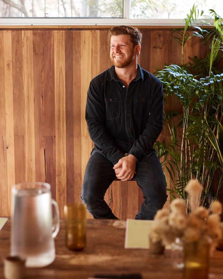 A smiling man in a dark shirt sits on a stool in front of a timber-panelled wall and palms, with the Bali table and dried flowers in the foreground.