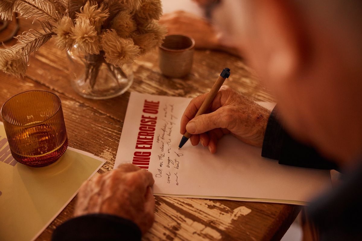 A hand writes on a sheet headed Writing Exercise One beside an amber glass and a vase of dried flowers on the Bali table.
