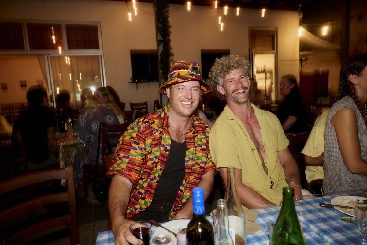 Two smiling guests pose at a blue-check table, one wearing a brightly patterned shirt and matching bucket hat.