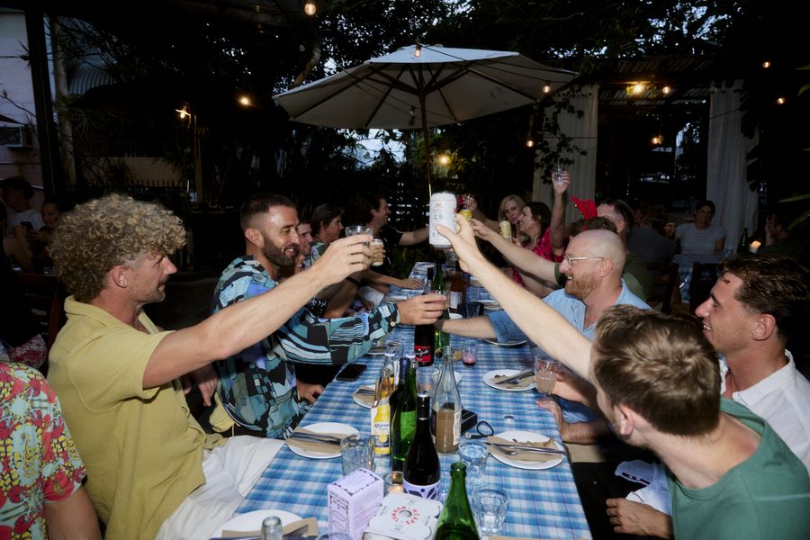 Guests smile and reach across a long blue-check table to toast beneath a large umbrella and string lights.