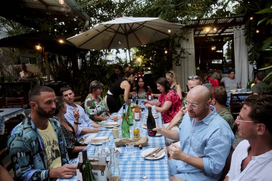 Guests gather around a long blue-check table beneath a large umbrella, surrounded by greenery and warm lights.