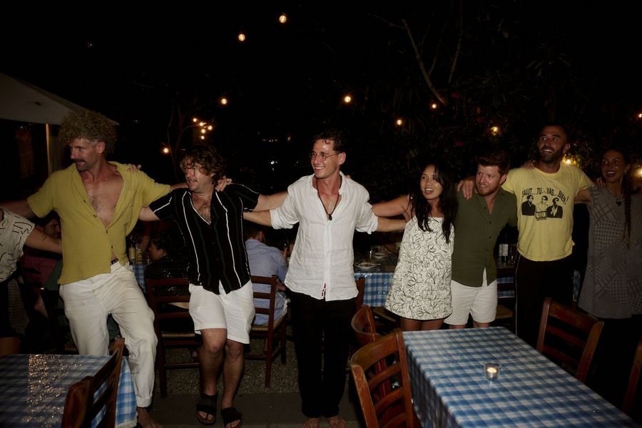 A line of smiling guests links arms and dances between blue-check dining tables under string lights.
