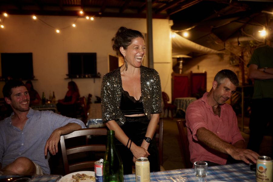 A guest in a sparkling dark jacket laughs while standing between two seated guests at the dinner table.